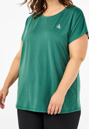 Frau trägt ein locker sitzendes grünes Kurzarmshirt mit einem kleinen dreieckigen Logo auf der Brust, kombiniert mit schwarzen Hosen.