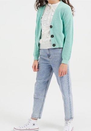 Kind trägt eine mintgrüne, geknöpfte Strickjacke, eine weiße Bluse mit Blumenmuster, hellblaue Jeans und weiße High-Top-Sneaker und steht vor einem weißen Hintergrund.