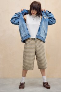 Denim jack, witte tanktop, geprinte shorts in beige en bruin, witte sokken en bruine moccasin-stijl schoenen. De achtergrond is licht hout.