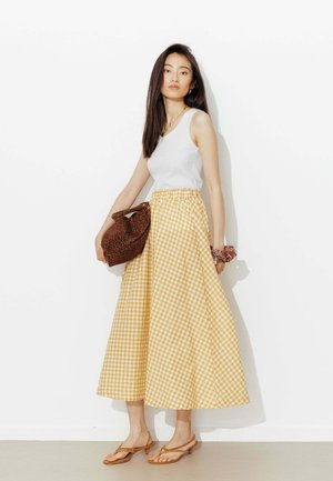 Femme debout contre un mur blanc portant un haut blanc sans manches, une jupe midi vichy jaune, des sandales marron, tenant un sac à imprimé léopard.