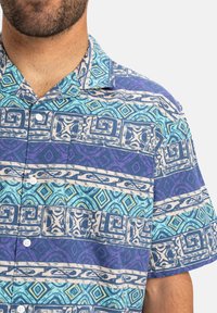 Camisa de botones de manga corta en azul y morado con patrones geométricos, con cuello y botones blancos en una tela texturizada.