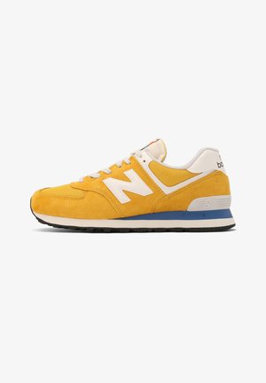 Żółty sneaker New Balance z białymi sznurowadłami, białym logo, niebiesko-białą podeszwą i czarnym bieżnikiem, widoczny od strony zewnętrznej.
