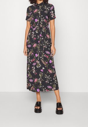 Kobieta w czarnej sukience midi z krótkimi rękawami, kołnierzykiem, zapinanej na guziki na przodzie, z kwiatowym wzorem w odcieniach różu i fioletu oraz paskiem wiązanym w talii.