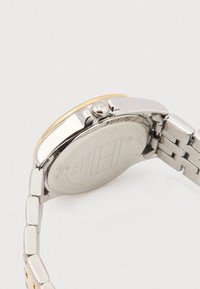 Stříbrné a zlaté kovové hodinky s kulatým ciferníkem, náramkem z propletených obdélníkových článků a výraznou korunou po straně. Na zadní straně se nachází logo značky.