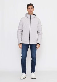 Giacca grigia chiaro con cerniera e cappuccio, caratterizzata da dettagli neri e tasche con cerniera. Indossata con jeans scuri e sneakers bianche. Tessuto liscio.