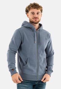 Lichtblauwe hoodie met rits, voorzien van een trekkoord in de capuchon, voorzakken, geribde boorden en een klein logo op de borst. Zachte stofstructuur.