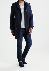 Abrigo trench azul marino con diseño de doble botonadura, hombreras y botones grandes, combinado con una camiseta gris y jeans oscuros.