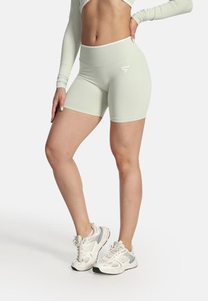 Donna che indossa pantaloncini da biker verde chiaro a vita alta e sneakers bianche, in piedi con una mano sui fianchi su sfondo bianco.