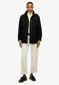 Chaqueta militar negra con múltiples bolsillos frontales y hombreras, llevada sobre un cuello de tortuga crema y pantalones blancos. Zapatos negros.