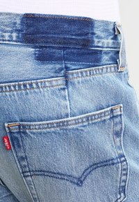 Close-up of light blue denim jeans showing back pocket with orange stitching and red Levi's fabric tag on side seam.