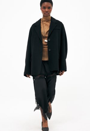 Kobieta idąca, ubrana w oversize'owy czarny płaszcz, brązową bluzkę z wysokim kołnierzem, czarną spódnicę wykończoną koronką, czarne szpilki i długi naszyjnik z zawieszką.