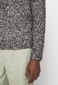 Suéter de punto en negro, gris y blanco con motas coloridas. Presenta un dobladillo y puños acanalados. Lleva puesto sobre pantalones verde claro.