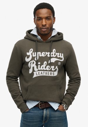 Olivkowa bluza z kapturem z białym nadrukiem "Superdry Riders Leathers", ze sznurkami ściągającymi, z kieszenią z przodu, noszona na jasnoniebieskiej koszuli z kołnierzykiem.