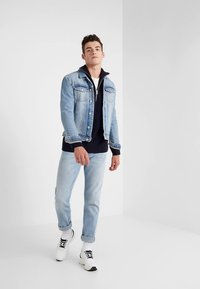 Giacca di denim azzurro chiaro sopra un maglione blu navy, abbinata a jeans azzurri chiari e scarpe da ginnastica bianche. La giacca presenta tasche e un colletto.