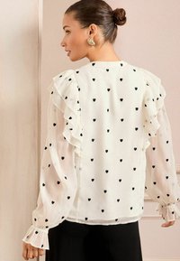 White blouse with black heart patterns, ruffled sleeves, and elastic cuffs. Lightweight, sheer fabric with a loose fit, styled for a feminine look.