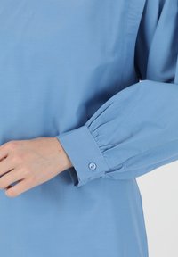 Camicia azzurro chiaro con vestibilità su misura, caratterizzata da maniche a sbuffo e polsini con bottoni. Realizzata in tessuto liscio con una leggera lucentezza.