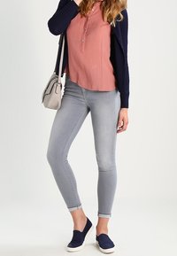 Vaqueros ajustados gris claro, cárdigan azul marino y una blusa rosa con botones. El calzado son zapatillas sin cordones azul oscuro. El bolso es pequeño, cruzado y de color gris claro.