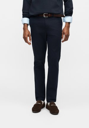 Férfi tengerészkék nadrágban, barna fonott övvel, barna mokaszinok fehér zoknival, világoskék ingujjak láthatók a tengerészkék pulóver alatt.