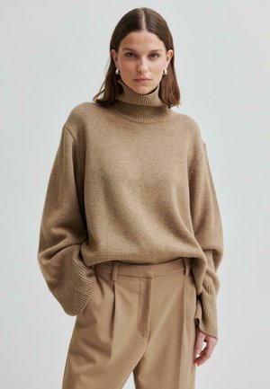 Kvinde med skulderlangt brunt hår iført en brun turtleneck-sweater og højtaljede beige bukser mod en ensfarvet baggrund.