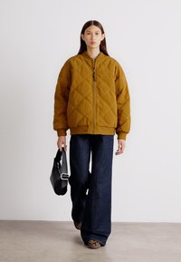 Gequilted mosterdjack met een ritssluiting aan de voorkant, geribbelde manchetten en een ontspannen pasvorm; gecombineerd met donkerblauwe flared jeans en een zwarte handtas.