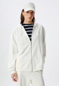 Felpa bianca con zip realizzata in un tessuto morbido, dotata di cappuccio e polsini a costine, abbinata a una camicia a righe blu e bianche sotto.