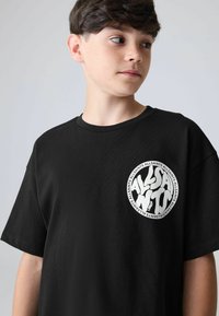 Poika, jolla on lyhyet tummat hiukset ja musta t-paita, jossa on valkoinen pyöreä logo ja tyylitelty teksti "ALLSAINTS" rinnassa, katsoo oikealle.