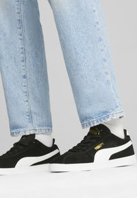 Fekete velúr sportcipők fehér részletekkel és arany logóval. Világoskék farmernadrággal viselve, frayed széllel és fehér zoknikkal.