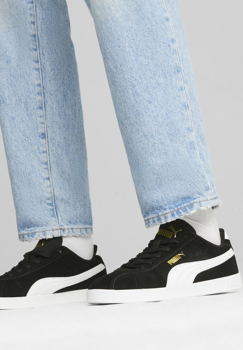 Fekete velúr sportcipők fehér részletekkel és arany logóval. Világoskék farmernadrággal viselve, frayed széllel és fehér zoknikkal.