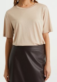 Beige tričko s krátkým rukávem a kulatým výstřihem, vyrobené z měkké tkaniny, spárované s tmavohnědou koženou sukní s hladkou texturou.