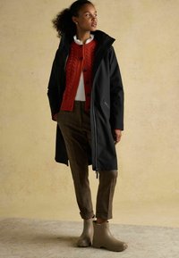 Palton lung negru peste un pulover roșu din tricot cu model cable-knit, pantaloni cu carouri și cizme negre până la glezne. Include fermoare și un guler înalt.