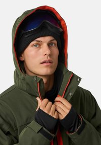 Olivgrüne Jacke mit orangefarbenem Innenfutter und schwarzen Akzenten. Sie verfügt über einen Reißverschluss, Druckknöpfe und Daumenlöcher an den Ärmeln. Eine schwarze Mütze und Skibrille werden getragen.