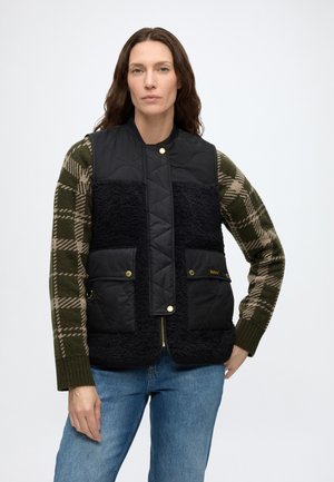 Frau trägt eine schwarze gesteppte und Fleeceweste über einem grünen und beigen karierten Pullover mit blauen Jeans, steht vor einem schlichten Hintergrund.