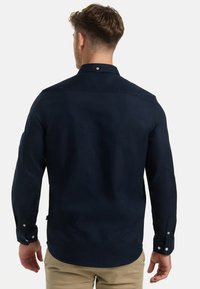 Mies nähtynä takaa, pukeutuneena tummansiniseen pitkähihainen paita ja beigeihin housuihin, yksinkertaista vaaleaa taustaa vasten.