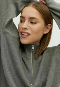 Grijze zip-up trui met ribbelstructuur en hoge kraag. Voorzien van een zilveren rits; de stof lijkt zacht en iets rekbaar.