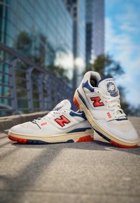 Baskets blanches avec accents rouges et bleus, matériaux en mesh et cuir, logo "N" bien visible, et semelles texturées avec des détails orange vif.