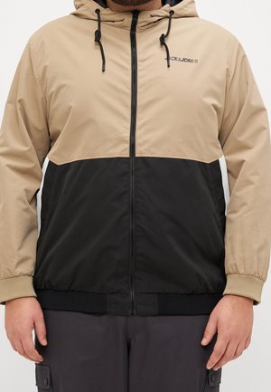 Mężczyzna w beżowo-czarnej kurtce z kapturem zapinanej na zamek, z logo Jack & Jones na piersi i elastycznymi mankietami.