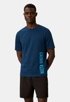 Uomo che indossa una t-shirt Calvin Klein blu navy con logo verticale sul lato e pantaloncini neri, in piedi con le mani dietro la schiena.