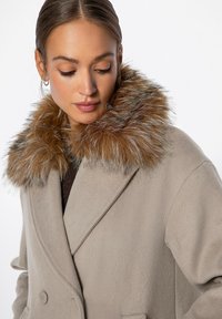 Beige doppeltknöpfiger Mantel mit einem plüschigen braunen Kunstpelzkragen und weicher Textur, ausgestattet mit einer sichtbaren Knopfleiste und tailliertem Design.