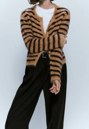 Personne portant un cardigan rayé marron et noir duveteux, une chemise blanche, un pantalon noir ample et une ceinture noire, debout, les bras croisés.