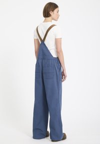 Blauwe katoenen salopettes met wijde pijpen, bruine corduroy schouderbanden en twee achterzakken. Het ontwerp heeft een relaxte pasvorm en minimale details.