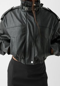 Veste noire en simili cuir courte avec col montant, fermeture à pression et ourlet élastique. Présente des accents métalliques et des épaules structurées.