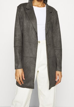 Osoba ubrana w ciemnoszary, długi żakiet na białą koszulę i białe spodnie z wysokim stanem, stojąca z ramionami swobodnie opuszczonymi wzdłuż ciała.