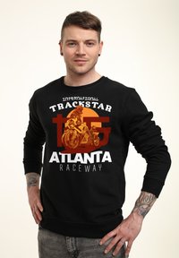 Svart hoodie med texten "International Trackstar" och "Atlanta Raceway" tryckt i vitt och orange. Har en motorcykelgrafik. Tillverkad av bomullsmaterial.