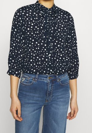 Kobieta nosząca granatową bluzkę w białe kropki z wiązaniem przy dekolcie, połączoną z wysokim stanem niebieskich dżinsów.