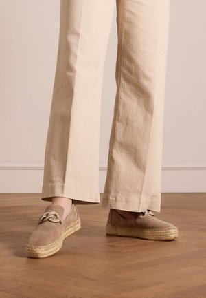 Beige suede espadrilles with a woven jute sole, featuring a decorative bow on the side. Paired with loose-fitting beige pants.