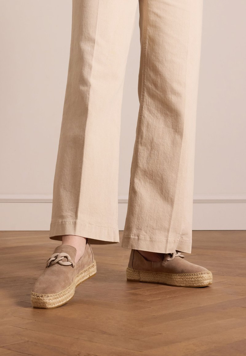 Beige suède espadrilles met een geweven jute zool, met een decoratieve strik aan de zijkant. Gecombineerd met een losse beige broek.
