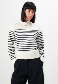 Maglione crema con strisce blu, collo a imbuto, tre bottoni al colletto e polsini a coste. Abbinato a pantaloni scuri e larghi.