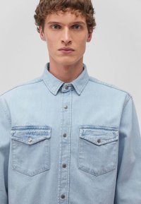 Camicia in denim azzurro chiaro con colletto, chiusura frontale a bottoni e due tasche sul petto. Presenta una texture leggermente sbiadita e bottoni in metallo argentato.