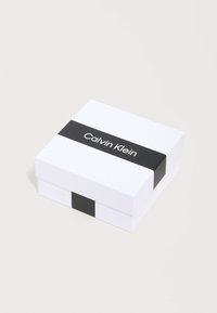 Λευκό τετράγωνο κουτί με μαύρη ταινία που φέρει το κείμενο «Calvin Klein», τοποθετημένο σε ανοιχτό γκρι επιφάνεια.