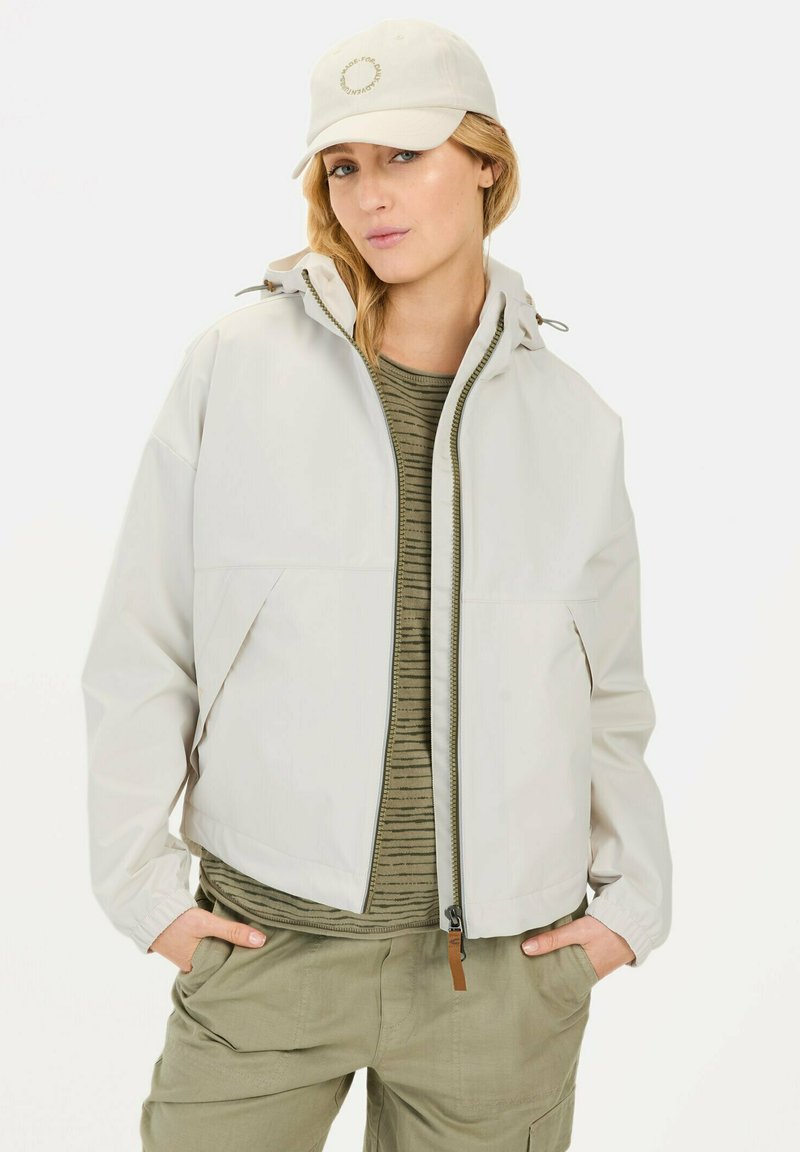 Lichtbeige geïsoleerde jas met een capuchon, ritssluiting en voorzakken, gelaagd over een groen gestreept shirt, gecombineerd met een beige pet.